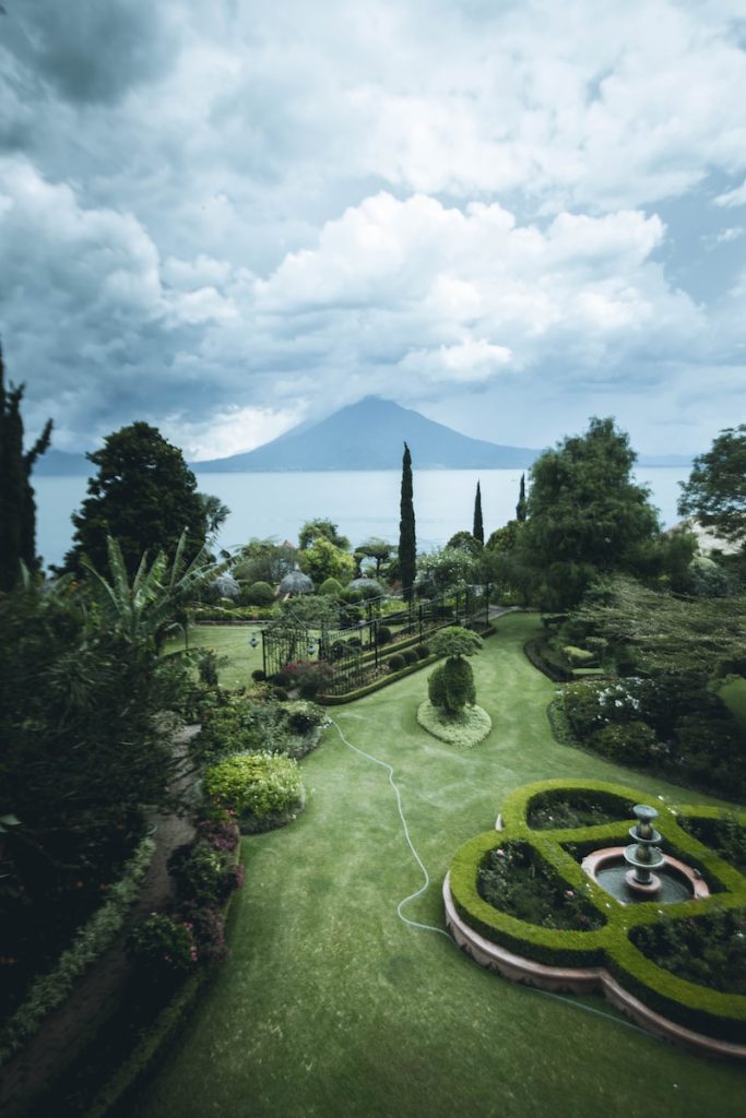 Scenic garden with hedges and fountain, overlooking a tranquil lake and distant mountain under a cloudy sky